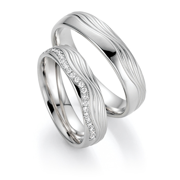 Zwei silberne Eheringe, einer davon mit einer Wellenlinie aus kleinen Diamanten, sind elegant auf weißem Hintergrund zu sehen - perfekt für Paare, die das Hochzeitshaus Konstanz für ihren besonderen Tag besuchen.