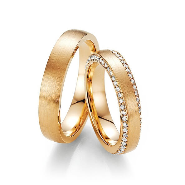 Zwei goldene Trauringe, einer schlicht und einer mit einer Reihe kleiner Diamanten, elegant in Szene gesetzt auf weißem Grund - ein zeitloses Symbol für Paare, die Brautmode und Herrenanzüge im Hochzeitshaus entdecken.