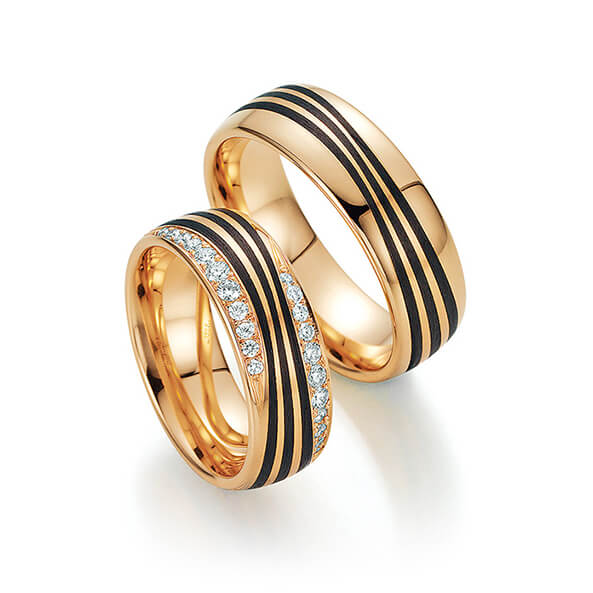 Zwei Goldringe mit schwarzen Streifen, einer davon mit einer Reihe kleiner Diamanten in der Mitte. Perfekt für Paare, die zeitlose Eleganz suchen. Erhältlich im Hochzeitshaus Konstanz neben den exquisiten Brautmode-Kollektionen.