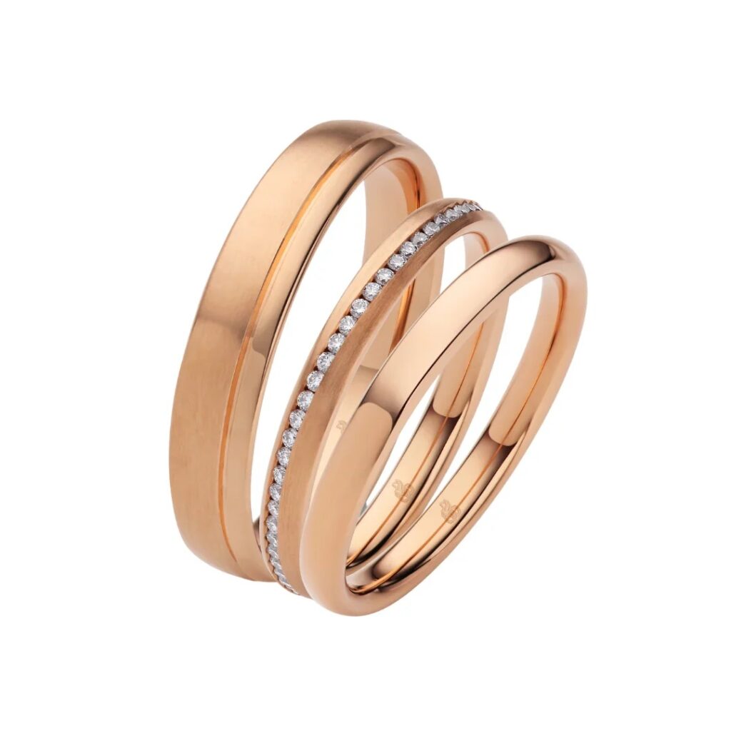 Drei Ringe aus Roségold, einer davon mit kleinen Diamanten, die auf weißem Grund aneinandergereiht sind.