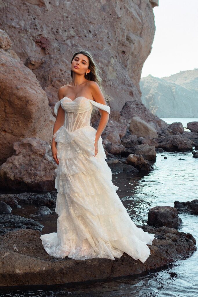 Eine Frau in einem weißen, schulterfreien Brautkleid steht auf Felsen am Meer, im Hintergrund sind Klippen zu sehen.