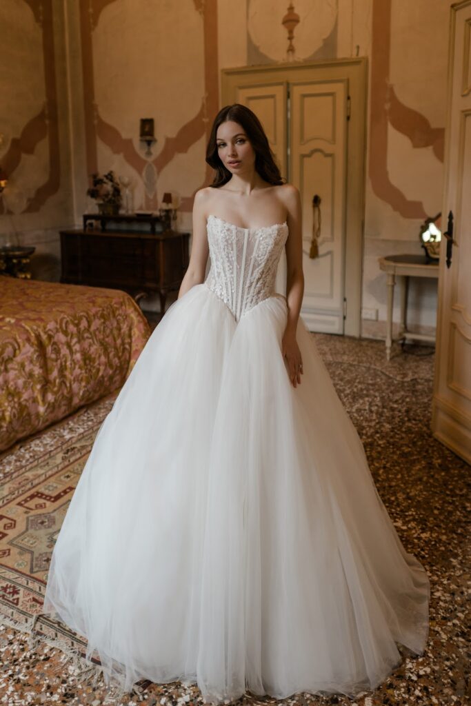 Eine Frau in einem trägerlosen weißen Brautkleid steht in einem eleganten Schlafzimmer im Vintage-Stil.