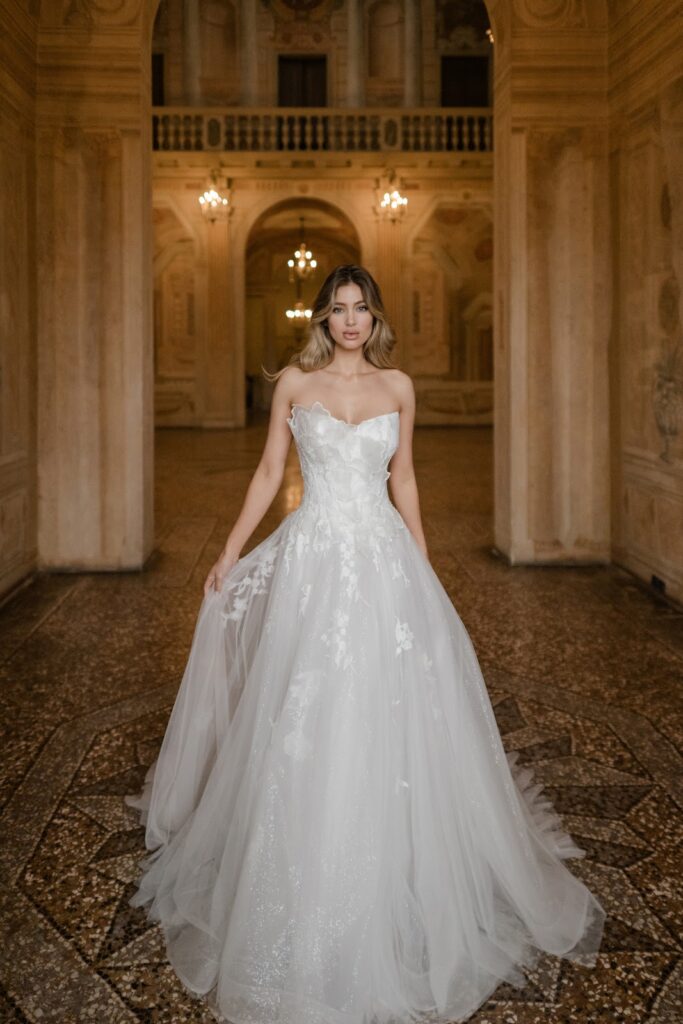 Eine Frau in einem trägerlosen weißen Brautkleid steht in einem prunkvollen, eleganten Saal mit warmer Beleuchtung.