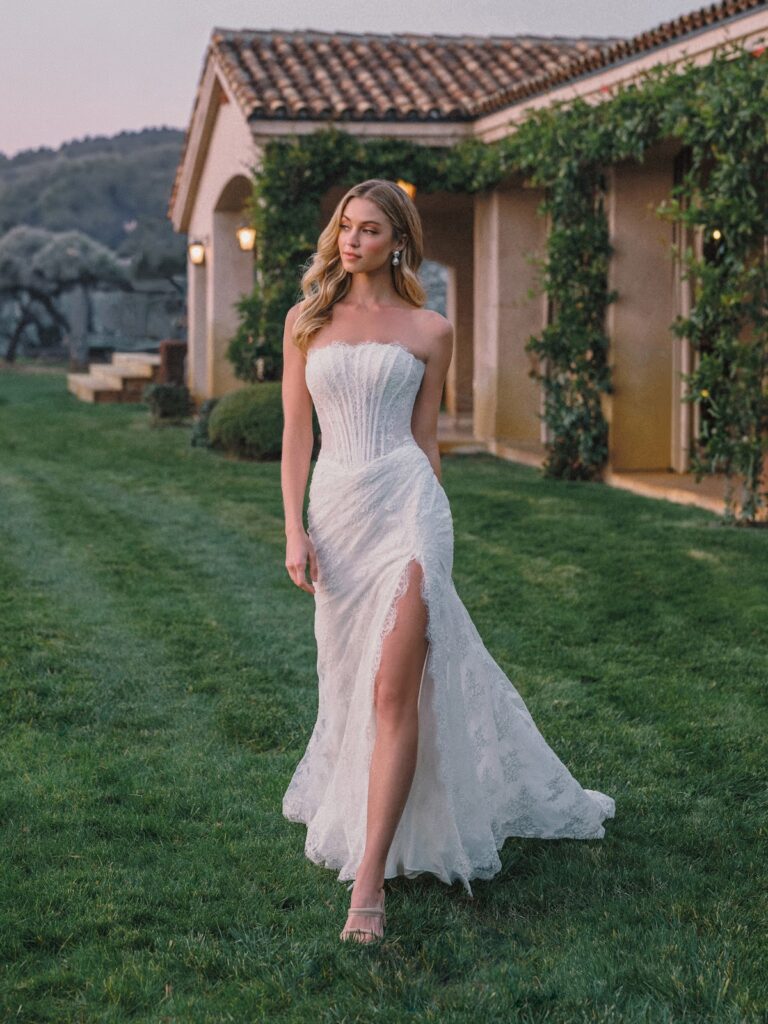 Eine Frau in einem trägerlosen weißen Brautkleid mit einem oberschenkellangen Schlitz, die bei Sonnenuntergang auf dem Rasen neben einer Villa spaziert.