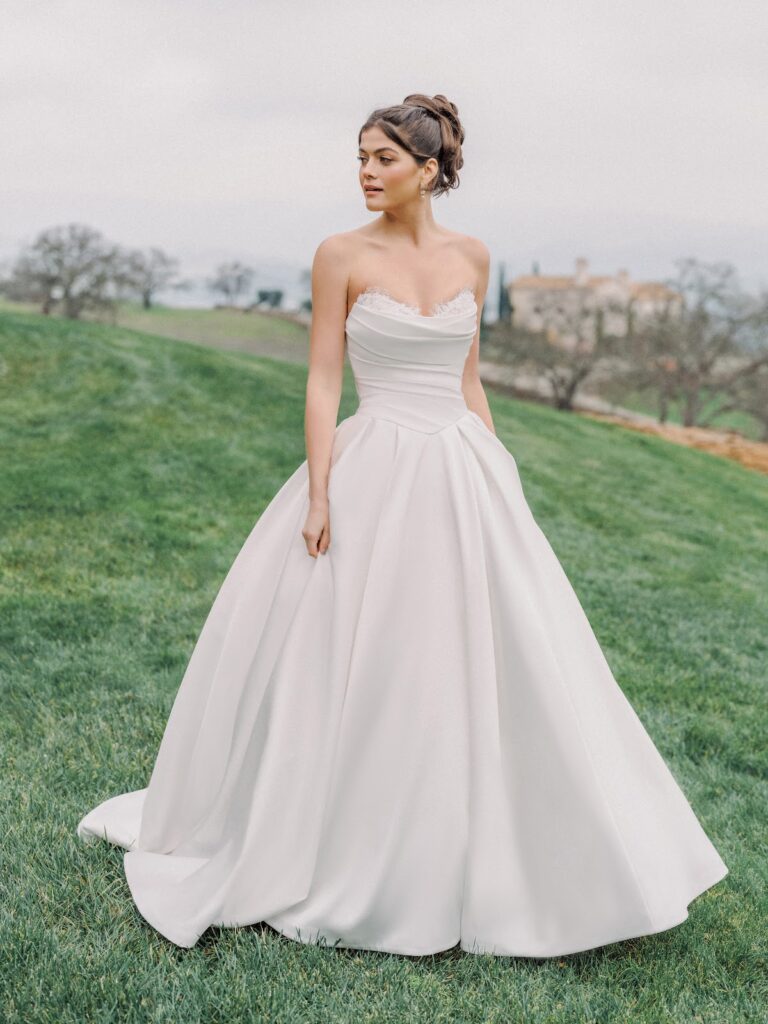 Eine Frau in einem trägerlosen weißen Brautkleid steht auf grünem Rasen, im Hintergrund sind Bäume und ein Gebäude zu sehen.