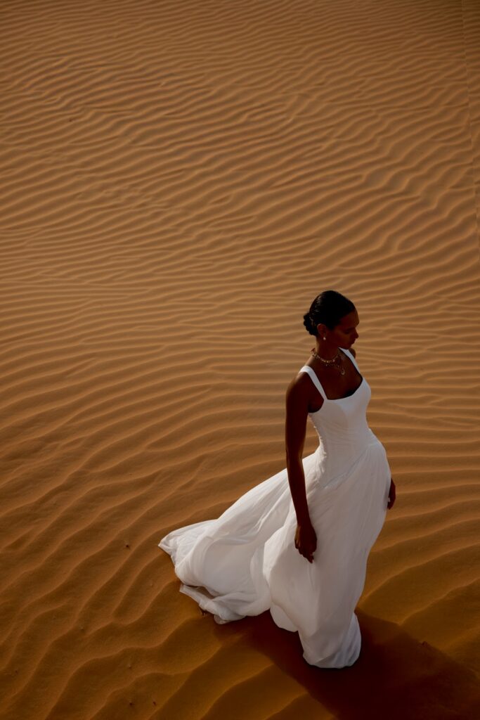 Eine Frau in einem weißen Kleid geht allein über wellige Sanddünen im warmen Licht.