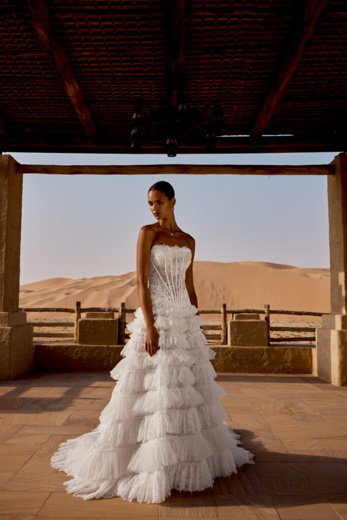 Eine Frau in einem trägerlosen, weißem Kleid mit Rüschen steht unter einem Holzdach vor Sanddünen in der Wüste.