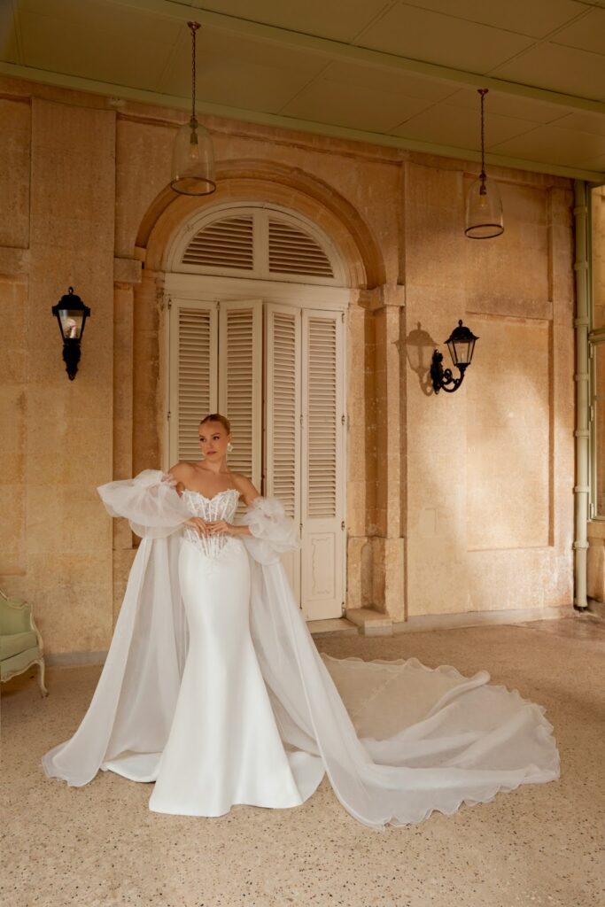 Eine Braut in einem eleganten weißen Kleid mit transparenten Ärmeln steht vor einem großen Torbogen.