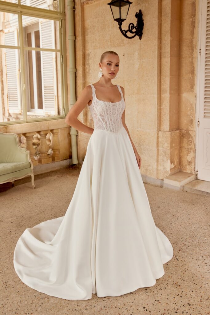 Eine Frau in einem eleganten weißen Brautkleid steht in einem Raum in der Nähe eines Fensters, im Hintergrund ist eine klassische Einrichtung zu sehen.
