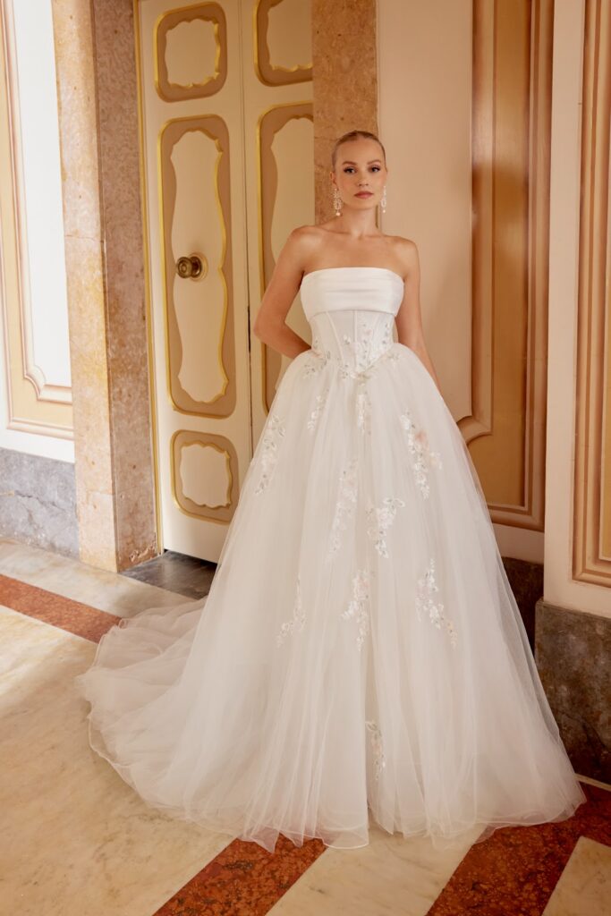 Eine Frau in einem trägerlosen weißen Ballkleid mit floralen Details steht in einem eleganten, reich verzierten Flur.