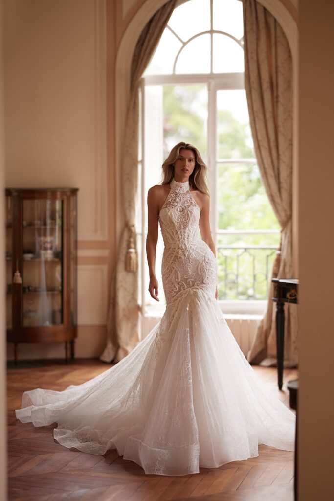Eine Frau in einem brautkleid aus Spitze im Meerjungfrauen-Stil steht in einem eleganten Raum mit hohen Fenstern und wallenden Vorhängen.