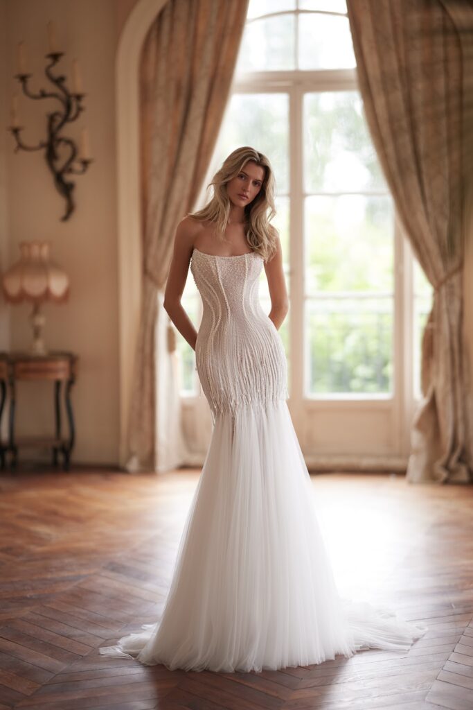 Eine Frau in einem trägerlosen, weißen Brautkleid im Meerjungfrauenstil steht in einem eleganten Raum mit großen Fenstern.