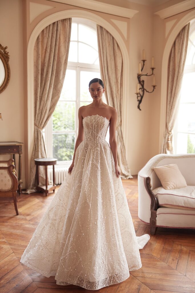 Eine Frau in einem trägerlosen, strahlend weißen Abendkleid steht in einem eleganten Raum mit hohen Fenstern und klassischer Einrichtung.