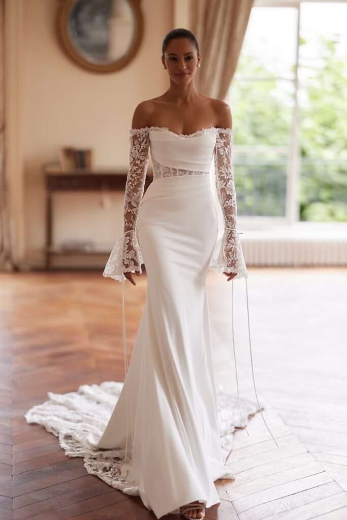 Eine Frau in einem eleganten weißen Brautkleid mit Spitzenärmeln steht in einem sonnendurchfluteten Raum mit Holzboden.