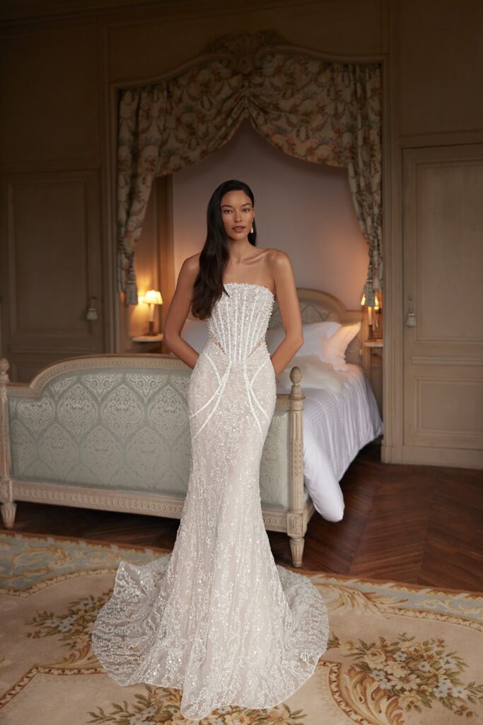 Eine Frau in einem trägerlosen, mit Perlen bestickten weißen Abendkleid steht in einem eleganten Schlafzimmer im Vintage-Stil mit einem Himmelbett.